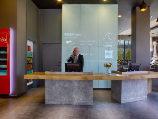 A professional receptionist assisting guests at the modern reception desk of Ibis Hotel Tuzla, Istanbul, with digital projections displaying hotel information.