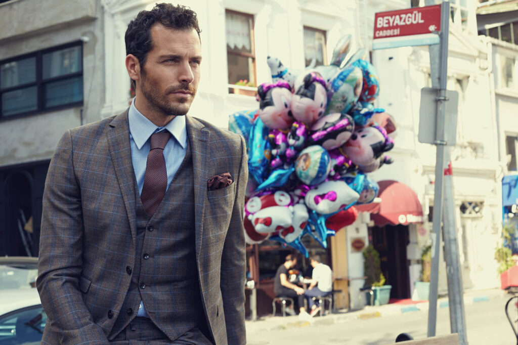A stylish man in a brown plaid three-piece suit, blue shirt, and brown tie from the Kigili Autumn Winter Menswear collection, posing in Beyoglu with a backdrop of colorful balloons.