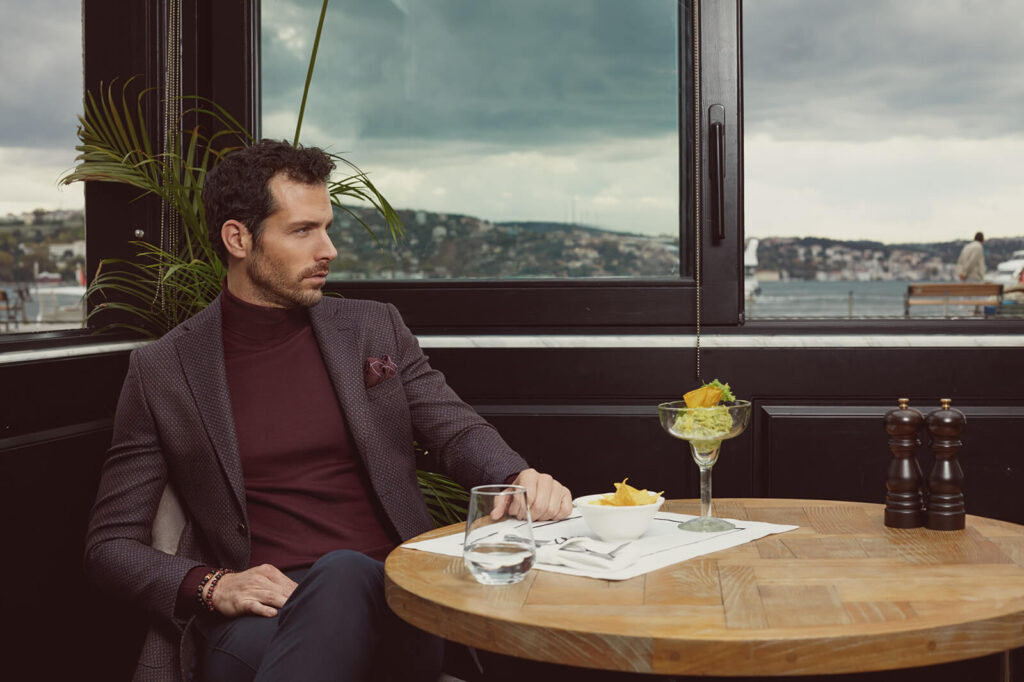 A stylish man in a burgundy turtleneck and dark blazer sitting at a restaurant with a Bosphorus view, part of Kiğılı&rsquo;s autumn-winter menswear collection.
