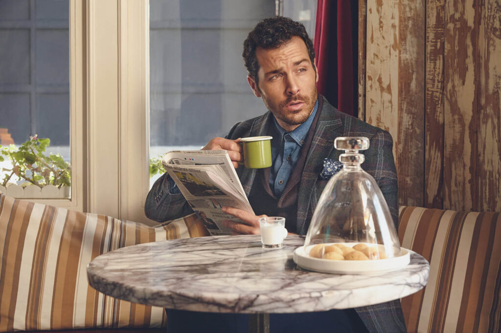A stylish man in a plaid blazer and blue shirt enjoying a coffee while reading a newspaper in a vintage caf&eacute;, part of Kiğılı&rsquo;s autumn-winter fashion campaign.