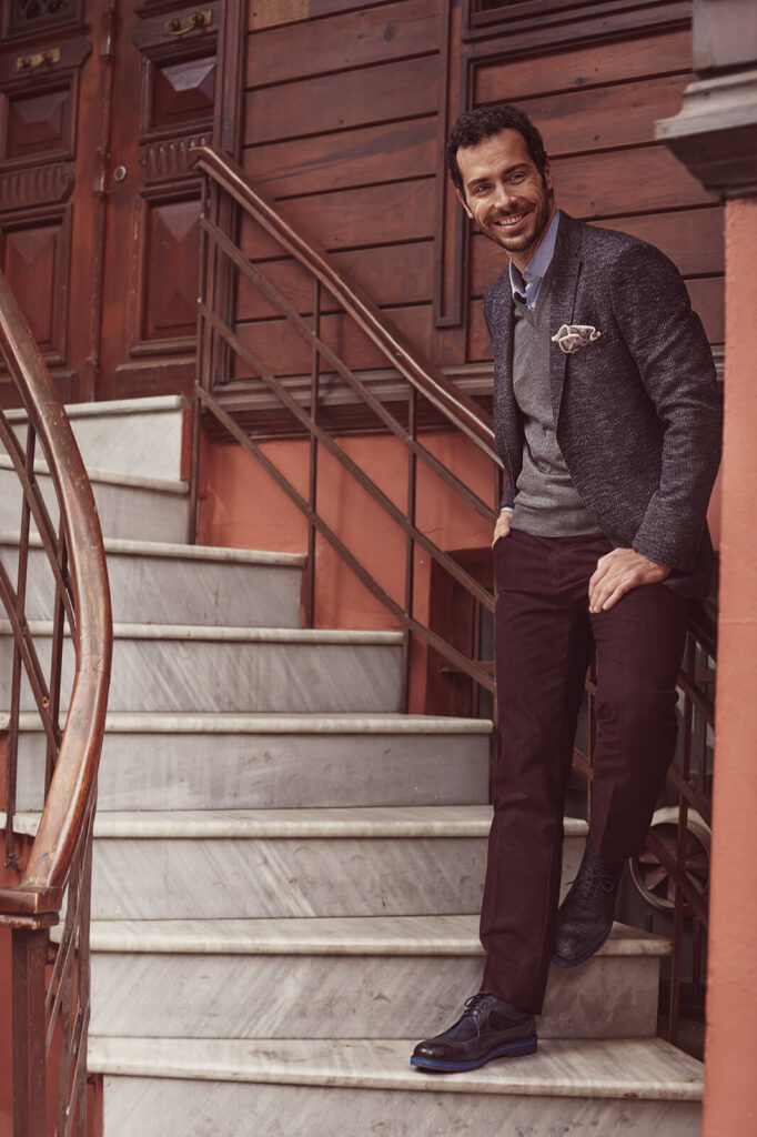 A stylish man in a dark blazer, gray sweater, and burgundy trousers smiling while stepping down marble stairs, part of Kiğılı&rsquo;s autumn-winter menswear collection.