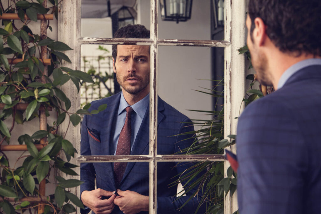 A stylish man in a navy plaid suit and brown tie looking at his reflection in an old window mirror, part of Kiğılı&rsquo;s autumn-winter menswear collection.