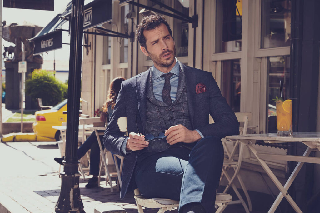 A stylish man in a navy-blue blazer, plaid vest, and tie sitting at an outdoor caf&eacute;, holding sunglasses, part of Kiğılı&rsquo;s autumn-winter menswear collection.