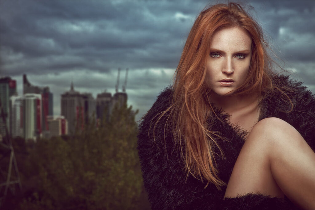 A fierce red-haired model in a black fur coat poses against a stormy city skyline.