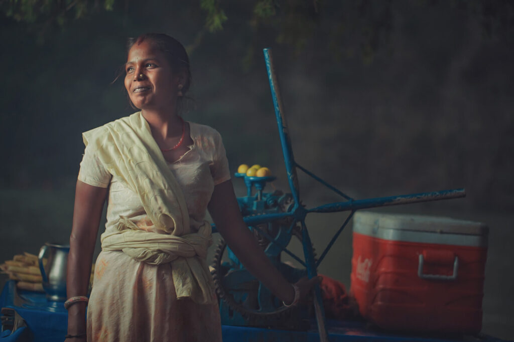 A smiling Indian woman selling sugarcane juice, standing beside her traditional juicing machine with fresh sugarcanes and lemons.